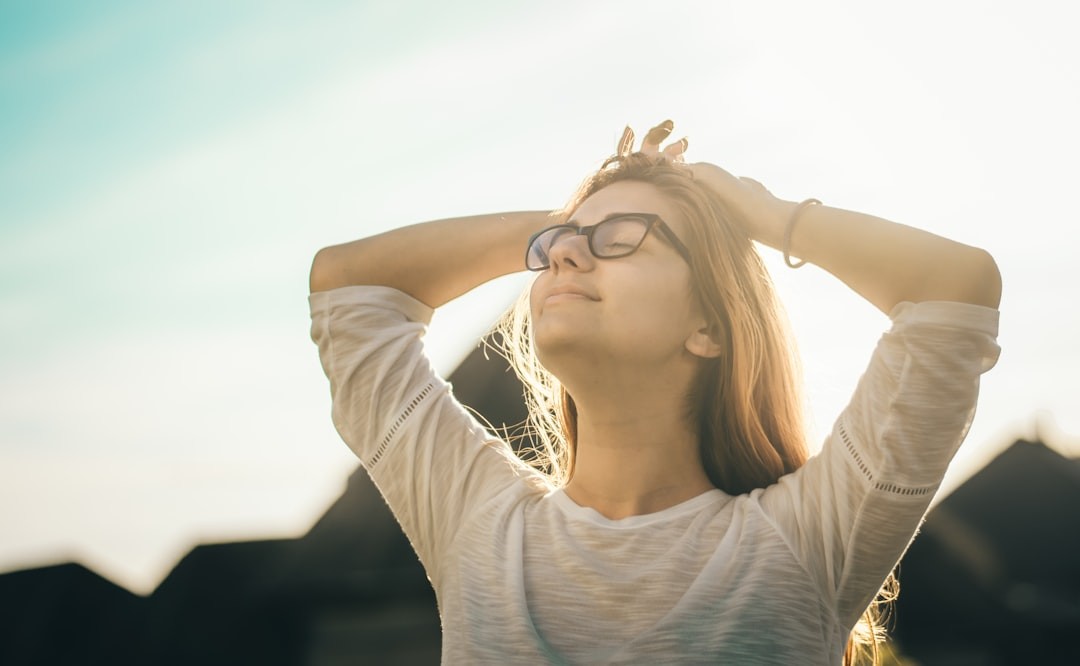 Person in a white shirt standing outdoors with hands on their head, basking in sunlight.